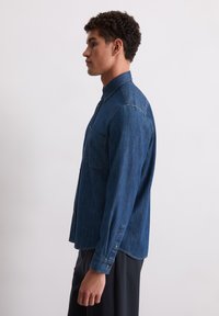Chemise en denim bleu foncé, avec un col boutonné, des manches longues et une poche poitrine; présente une texture lisse et une coupe décontractée.