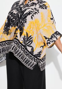 Blouse à manches courtes avec un motif floral noir et jaune sur fond beige, dotée d'un ourlet à motif contrastant.
