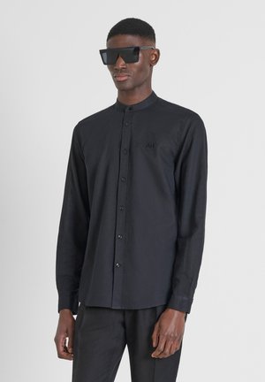 Schwarzes Langarmshirt mit Mandarin-Kragen, Knopfleiste und gesticktem Logo. Glattes Material und taillierte Passform, kombiniert mit großen schwarzen Sonnenbrillen.