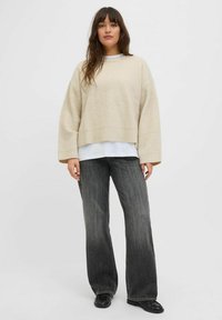 Suéter beige de canalé con mangas largas, combinado con jeans de pierna ancha gris y zapatos negros. El conjunto presenta un look en capas con una parte superior blanca por debajo.