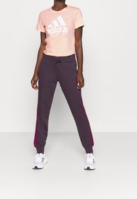 Camiseta de algodón rosa claro con logo blanco de Adidas, combinada con pantalones jogger gris oscuro con rayas fucsias en los lados y cintura elástica. Zapatillas blancas.