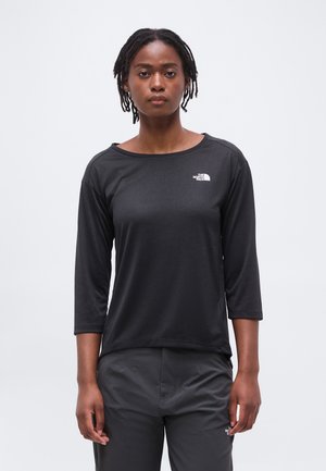 Γυναίκα με κοντά dreadlocks που φοράει μαύρο πουκάμισο με μανίκια τριών τετάρτων και σκούρο παντελόνι με λογότυπα The North Face, που στέκεται μπροστά σε λευκό φόντο.