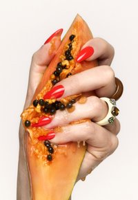 Kawałek papai jest wyciskany, odsłania pomarańczowy miąższ i czarne nasiona. Dłoń ozdobiona czerwonymi paznokciami i pierścionkami, teksturowana powierzchnia z sokiem.