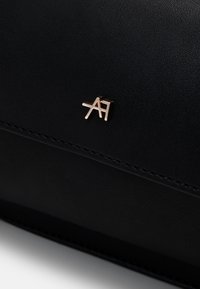 Sac en cuir noir avec un design cousu et un accent logo en ton doré. La texture est lisse avec une légère brillance et un matériel minimaliste.