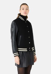 Zwarte varsity jack met een wollen lichaam, leren mouwen, gestreepte ribgebreide manchetten en een drukknoopsluiting met zijzakken. Geborduurd nummer '73' op de mouw.