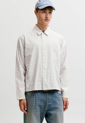 Junger Mann mit hellblauer Kappe, weiß kariertem Langarmhemd und blauen Jeans, stehend vor einem schlichten weißen Hintergrund.