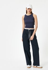 Camiseta sin mangas de rayas marinas, pantalones negros holgados con cordón blanco y gorra blanca. El modelo usa zapatos grises con rayas oscuras y claras.