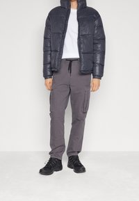 Gewatteerde marineblauwe pufferjas met ritssluiting en hoge kraag; wit T-shirt; grijze cargobroek met zakken aan de zijkant; zwarte veterschoenen.