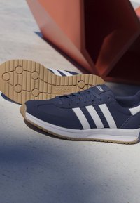 Zapatillas Adidas azul marino con rayas blancas, suela de goma marrón claro y diseño con cordones sobre una superficie clara junto a una estructura roja geométrica.