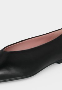 Ballerine plate en cuir noir avec un bout pointu et une doublure intérieure rose douce, présentée sur un fond uni gris clair.