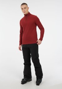 Maglione rosso a maniche lunghe con zip e colletto alto, abbinato a pantaloni neri larghi e scarponi da sci, su uno sfondo bianco puro.
