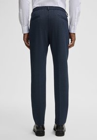 Ciemnogranatowe spodnie z teksturowanej tkaniny, z elastycznym paskiem w talii i dwoma tylnymi kieszeniami; w połączeniu z czarnymi butami. Widok od tyłu.