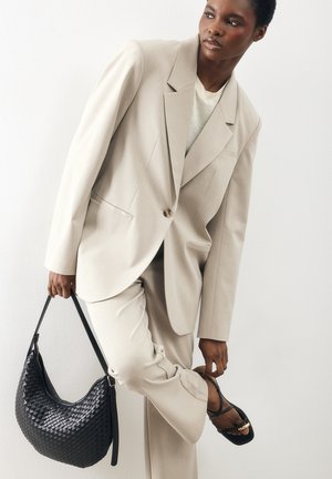 Kvinde i beige jakkesæt, der justerer sort sandal med remme, mens hun holder en sort flettet håndtaske mod en hvid baggrund.