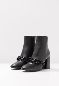 Botas de cuero negro hasta el tobillo con punta afilada, tacón grueso y un adorno de cadena negra decorativa alrededor del tobillo. Cierre de cremallera en el lateral.