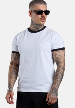 Man with tattoos wearing black sunglasses, white T-shirt with black trim, and black pants, looking to the side against a gray background.