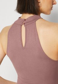 Bezramenný top v mauve tkanině s vysokým límcem, na zadní straně s klíčovou dírkou, která je zajištěna dvěma knoflíky. Hladká textura s minimálními švy.