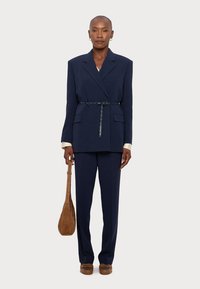 Ναυτικό μπλε σακάκι κομμένο με ζώνη, αξεσουάρ από ασημί υλικό, παντελόνι σε ταιριαστή απόχρωση, και ανοιχτόχρωμο πουκάμισο; μια καφέ τσάντα ώμου.