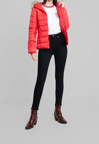 Rode gewatteerde jas met capuchon met bontkraag, gedragen over een wit t-shirt en zwarte skinny jeans met hoge taille, gecombineerd met glanzende bruine enkellaarzen.