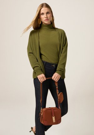 Olivgrüner Strickcardigan über einem passenden Rollkragenpullover, kombiniert mit dunklen Jeans. Eine kleine braune Handtasche mit goldenen Akzenten und einem Tragriemen.