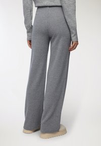 Persona che indossa pantaloni a gamba larga a vita alta in maglia grigia e scarpe con plateau beige chiaro, in piedi contro uno sfondo semplice.