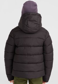 Zwarte pufferjack met horizontale quilting, een nauwsluitende capuchon en elastische manchetten. Draagt een lichtpaarse gebreide beanie.