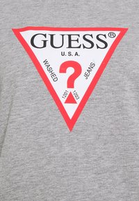 Šedé bavlněné tričko s červeným trojúhelníkovým logem s texty "GUESS" a "U.S.A.", centrálním otazníkem a okolními klíčovými slovy v černé barvě.