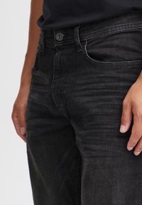 Czarne jeansy z denimu z widocznymi przetarciami, o wąskim kroju, standardowym układzie kieszeni i kontrastującym przeszyciem. Zapięcie na guziki z przodu.