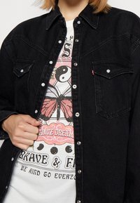 Musta denimi-paitapusero, jossa on napsupainikkeet, kaksi rintataskua ja terävä kaulus, kerrostettuna värikkäillä kuvioilla varustetun valkoisen t-paidan päälle.