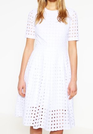 Femme portant une robe blanche longueur genou avec des manches courtes et un motif circulaire perforé sur toute la matière.