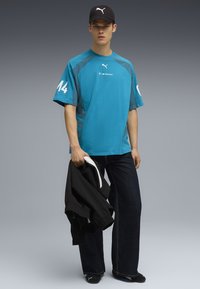 Turkos oversized t-shirt med blå accenter, med "14" på ärmen. Svart keps, mörka jeans och en svart jacka i handen.