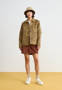 Kožený kabát s leopardím potiskem v béžové a hnědé barvě, nošený přes sukně barvy rez. Bílá teniska a béžový klobouk s širokým krempou dotvářejí vzhled.