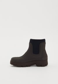 Botín de lluvia negro grueso hasta el tobillo con paneles laterales elásticos y suela gruesa en fondo blanco.