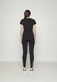 Schwarzes T-Shirt mit Rundhalsausschnitt und kurzen Ärmeln, kombiniert mit schwarzen Leggings. Die Leggings haben einen weißen Akzent. Das Model trägt schwarz-weiße Sneakers.