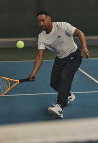 White athletic t-shirt with black logo, black pants, and white sneakers with black accents. Player hits a yellow tennis ball with an orange racket.