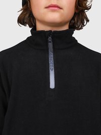 Primo piano di una persona che indossa un giaccone in pile nero con colletto alto con zip, mostrando un testo parziale sulla striscia della zip.