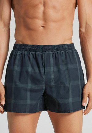 Mandlig overkrop iført mørkegrå ternede boxershorts med elastisk talje, stående mod en ensfarvet lys baggrund.