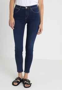 Mörkblå skinny jeans med hög midja, med kontrasterande sömmar och knappstängning fram. Kautat med svarta sandaler.