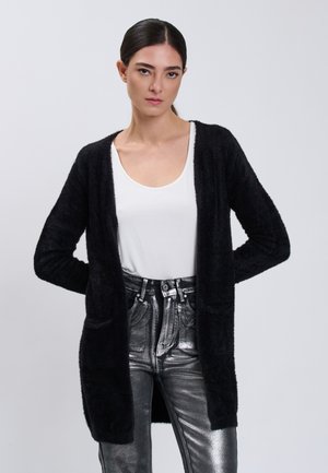 Mujer con cabello oscuro que lleva un cárdigan negro y peludo sobre una camiseta blanca y unos vaqueros metálicos plateados de talle alto sobre un fondo liso.
