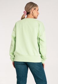 Jasnozielona bluza z luźnym krojem, ściągaczami przy mankietach i u dołu. Miękka tekstura z okrągłym dekoltem, noszona na ciemnoniebieskich dżinsach.
