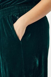 Groene fluwelen broek met een elastische tailleband, voorzien van een zijzak. De stof heeft een gladde, glanzende textuur en een diepe, rijke kleur.