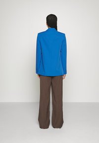 Blue blazer with a tailored design, peak lapel, and back yoke detail; paired with loose, brown trousers. Smooth texture and structured shape.