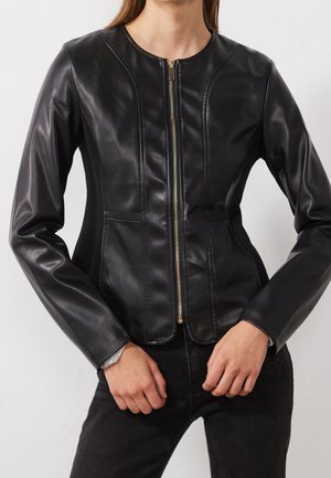 Femme portant une veste en cuir noire ajustée avec une fermeture éclair frontale, associée à un pantalon noir, mains près de la taille.