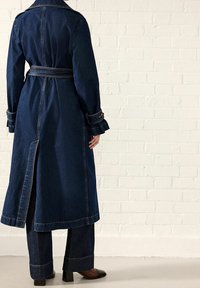 Persona vestida con un largo abrigo de mezclilla oscuro con cinturón, pantalones de mezclilla de pierna ancha a juego y zapatos de tacón marrones, de pie contra una pared de ladrillo blanco.