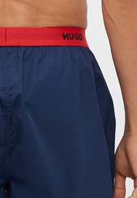 Shorts de bain bleu marine avec une ceinture élastique rouge arborant le logo "HUGO" en noir. Le tissu est lisse et léger.