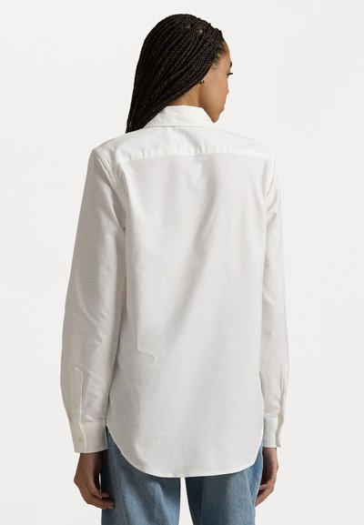 Hvit langermet skjorte med krage, med knapper ved mansjettene og rundet nederkant. Stoffet ser glatt ut med en svak glans.