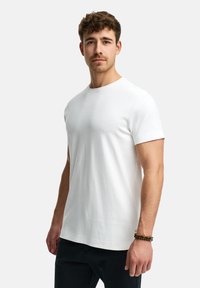 Weißes T-Shirt mit kurzen Ärmeln und Rundhalsausschnitt, aus glattem Stoff gefertigt. Enthält ein kleines Logotäfelchen am Saum. Begleitet von einem Armband.