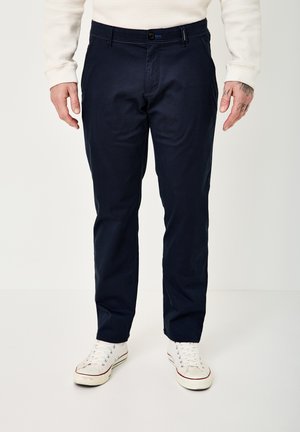 Mann trägt marineblaue gerade geschnittene Hosen, weiße High-Top-Sneaker und ein weißes strukturiertes langärmeliges Hemd, steht vor einem schlichten Hintergrund.