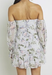 Robe florale dénudée d'épaule en tissu léger et transparent. Elle présente des roses pastel sur un fond clair, un ourlet volanté et des manches longues.