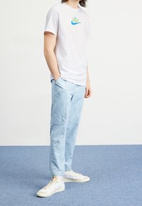Osoba ubrana w biały t-shirt z grafiką Nike, jasnoniebieskie spodnie i białe sneakersy stoi na niebieskim dywanie w tle białej ściany.