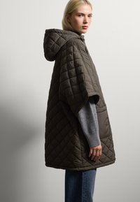 Poncho imbottito con cappuccio in verde oliva scuro, con maniche corte, una vestibilità rilassata e una texture morbida, abbinato a un maglione grigio e jeans.